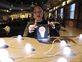 Person sitting at a table interacting with a light bulb box with a string of lit lightbulbs on the table